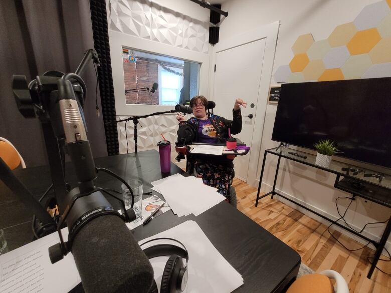 A woman sits in an audio studio in a wheelchair with a microphone in front of her. There's a studio table and another microphone in the forefront. 