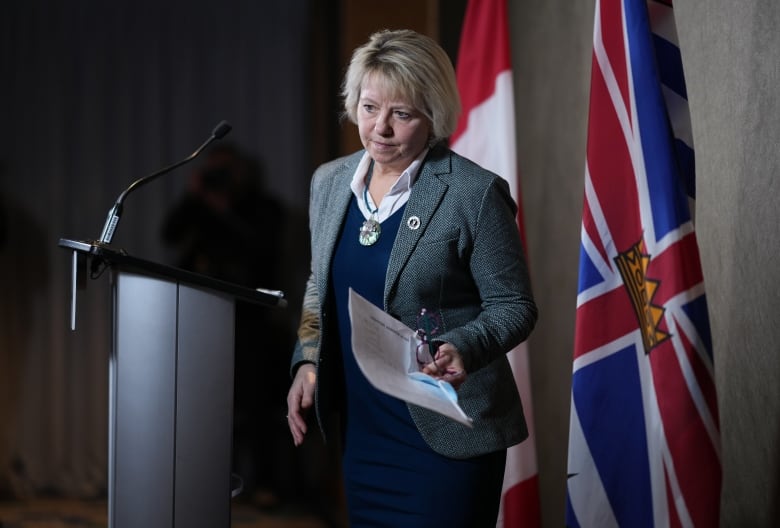 A woman with a blond bob haircut, wearing a grey blazer over a navy blue sweater steps away from a podium. In her left hand, she holds speaking notes and a pair of reading glasses.