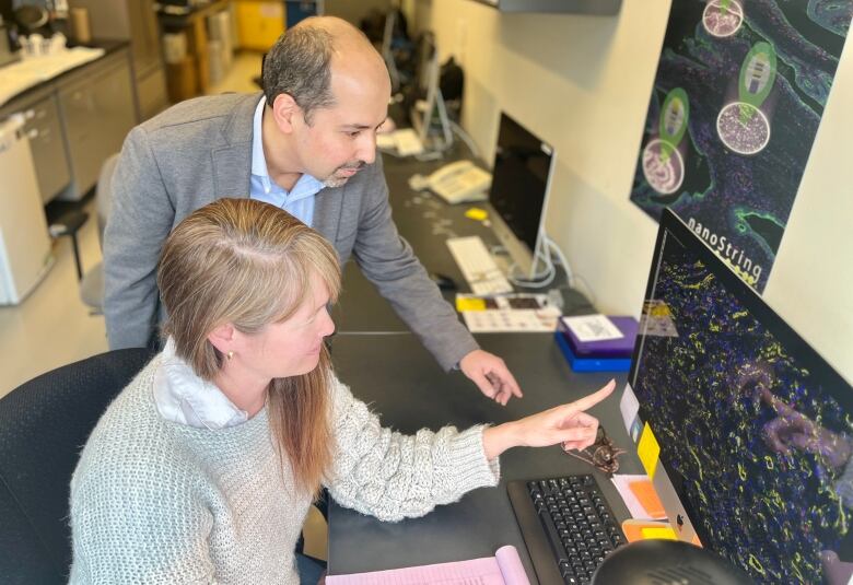 A man stands slightly above a woman seated in front of a computer screen, they are both looking at the screen while she point to the image of cancer cells displayed.