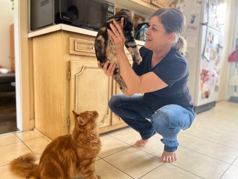 A woman crouches on the floor of her kitchen, holding up a grey and black cat, smiling at the cat.