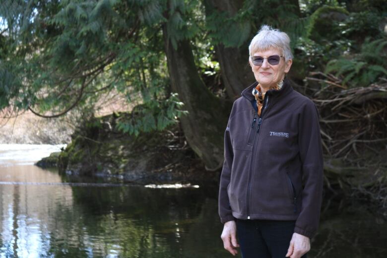 A woman wearing sunglasses and a fleece stands by a still river overhung by the branches of a large tree.