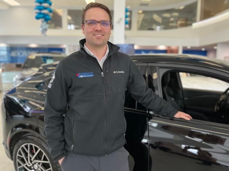 A worker at a car dealership stands beside a car for sale.