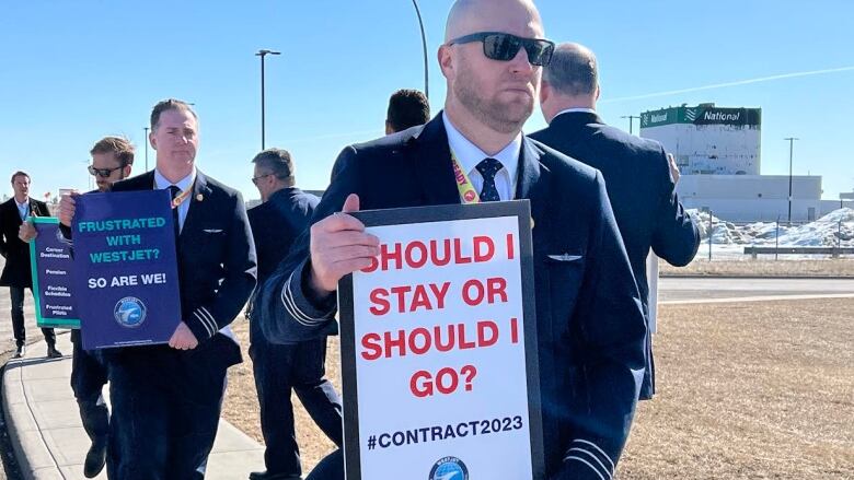 A bald man wearing sunglasses and a suit holds a sign that reads 'should I stay or should I go?.'