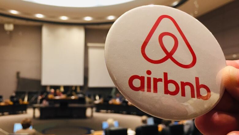 An Airbnb pin is held up against the backdrop of a city council chamber.