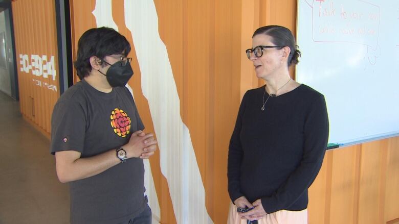 A man wearing a CBC shirt and a black facemask talks to a woman with black hair and wearing a black top.