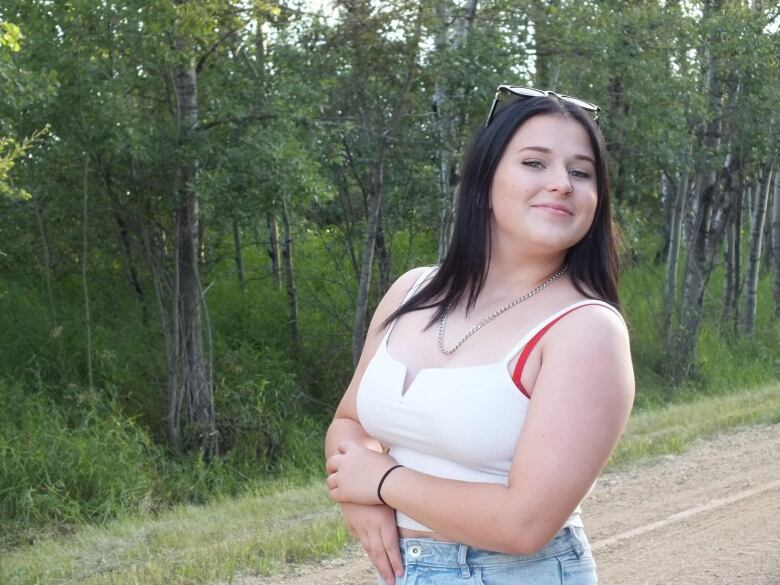 A teenage girl wearing a white tank top and light-coloured blue jeans stands near a line of leafy green trees.