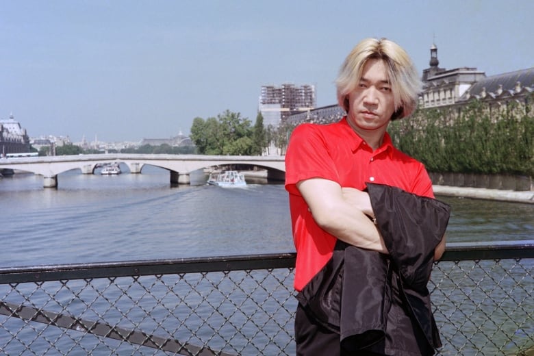 A man wearing a red shirt is pictured standing in front of a low fence with a canal in the background.