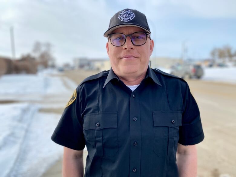 A clean-shaven white man is wearing large black-rimmed glasses, a black baseball cap and his firefighter uniform. He is standing outside, on the side of the street. It's an overcast day with snow on the ground.