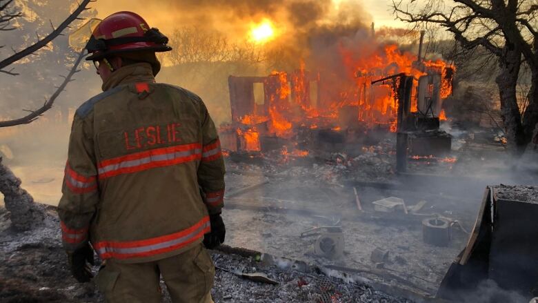 A firefighter in full gear stands in front of burning rubble. The rubble was once a house. There is smoke and ash all around. The morning sun shines a golden hue through plumes of smoke emanating from the fire. 