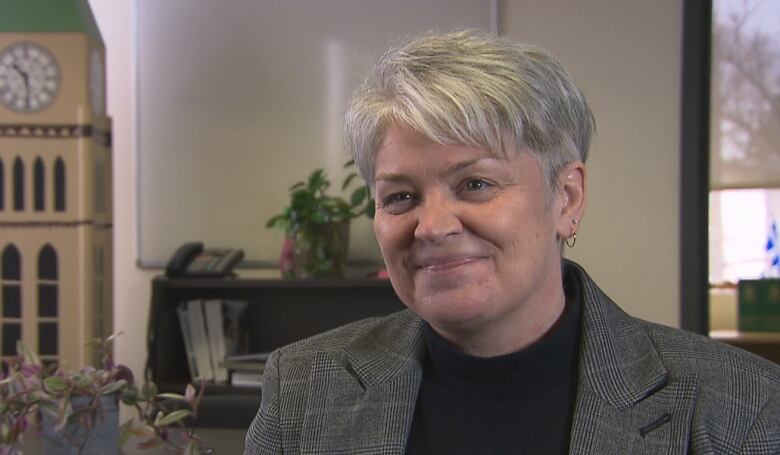 A woman with short grey hair is wearing a black turtleneck under a grey blazer. She's smiling while sitting in an office. Behind her is a replica clocktower, two plants, a bookshelf and a landline telephone.