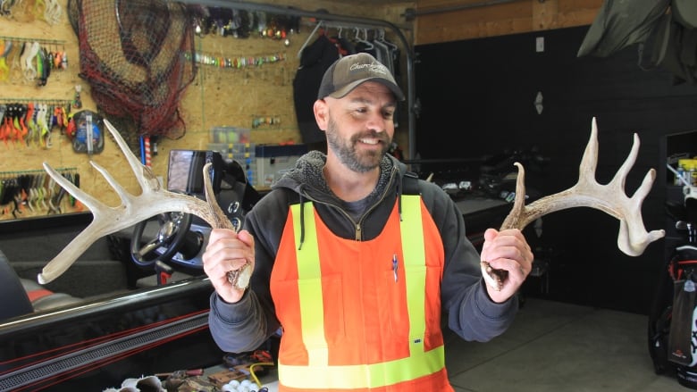 A man wearing orange overalls holds up two deer antlers. 