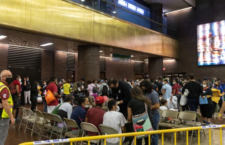 Large numbers of migrants wait in line and sit on chairs in a large room at a bus terminal.