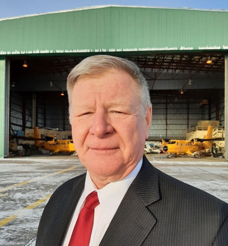 A man in a suit stands outside with a large airplane hangar visible behind him.