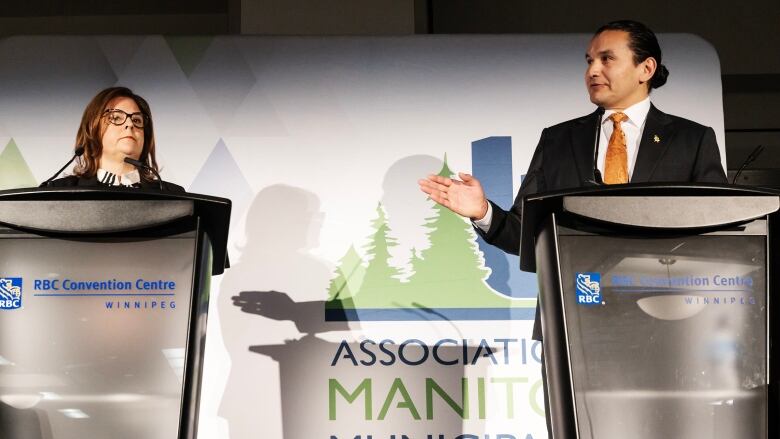 A man at a podium on the right gestures with his right hand toward a woman at a different podium on the left.