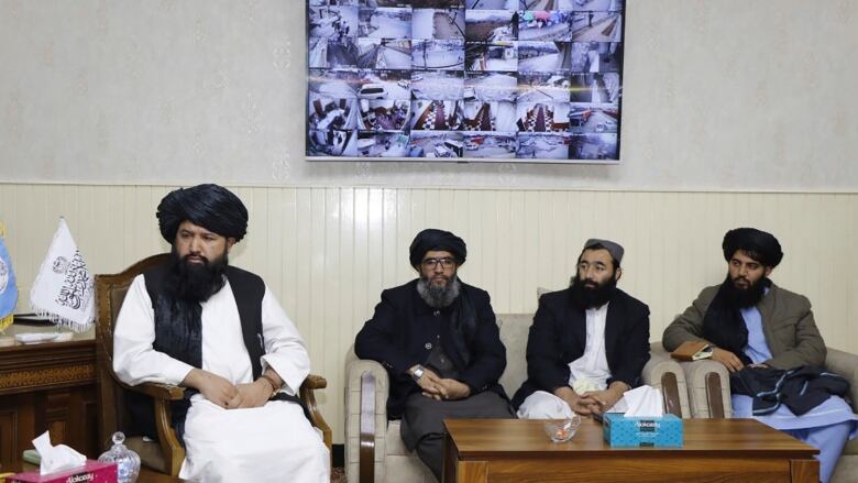 Four men sit in a room with a TV screen on the wall behind them.