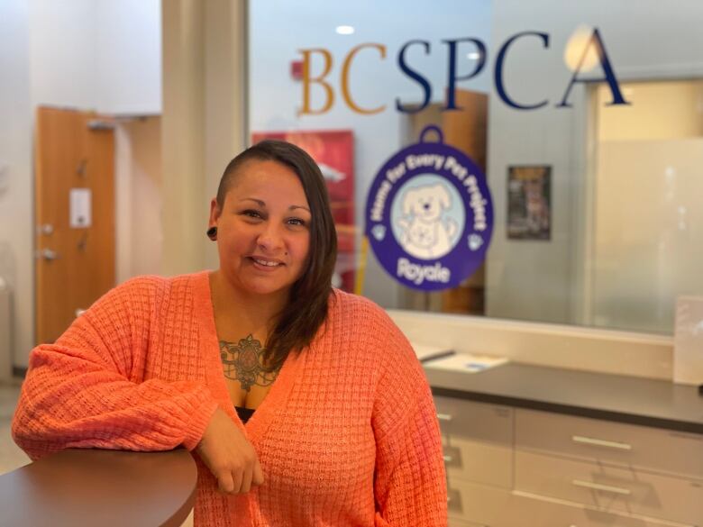 A woman in orange stands in front of a window with words that read BCSPCA.