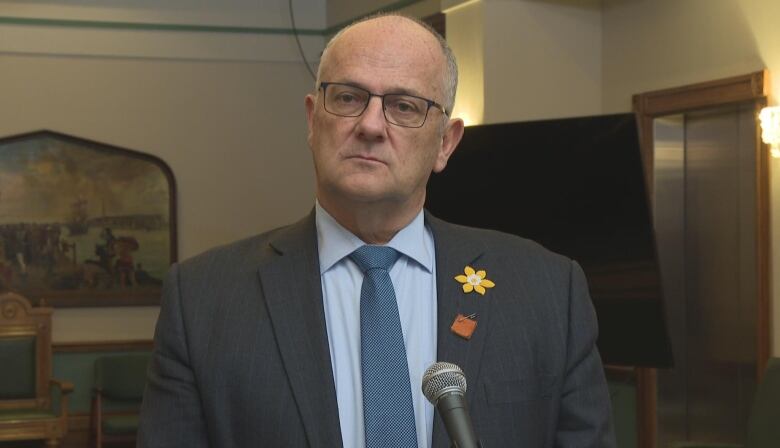 A man in glasses and a grey suit stands in front of a microphone.