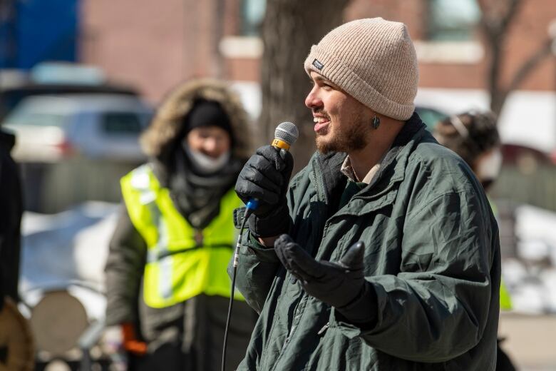 A person in a beige toque and a green coat speaks outside, holding a microphone.