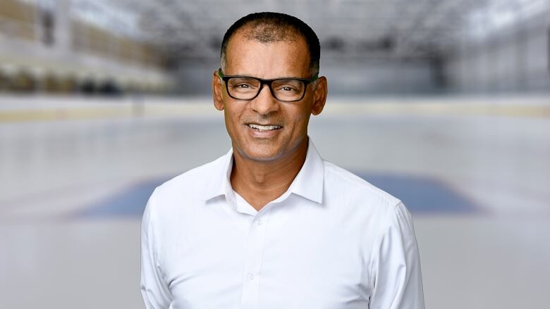 A man with glasses and black hair smiles at the camera in front of a hockey rink.