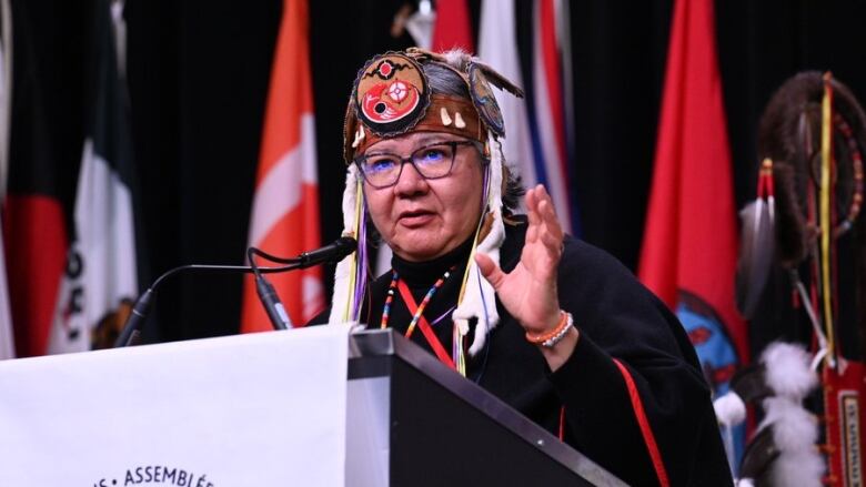 The national chief at a podium wearing an ornate headdress raises her hand as she speaks.