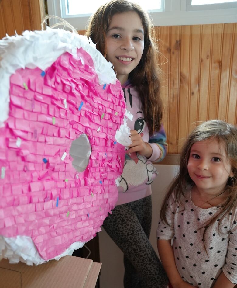 Two young girls stand side by side holding and white and pink pinata shaped like a frosted doughnut.