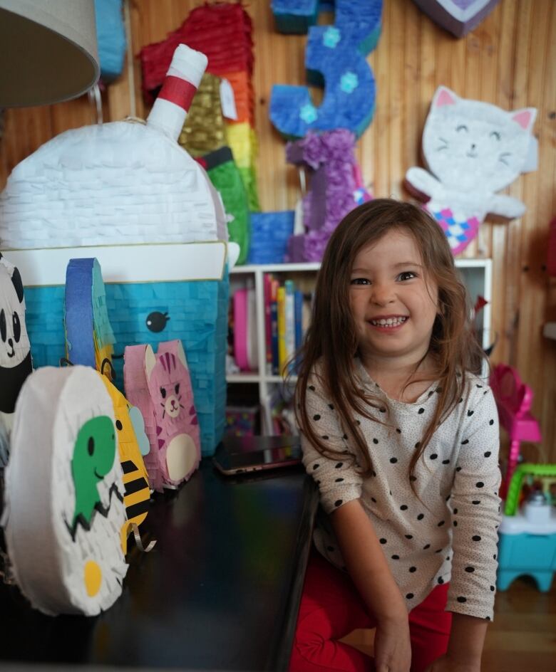 A little girl in white polka-dot shirt and red pants smiles standing next to a row of colourful pinatas. 