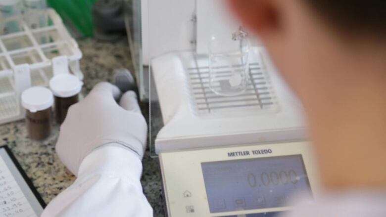 The back of a man's head and his hands, wearing white gloves, moving a small number of paint chips into a glass beaker on a scale.