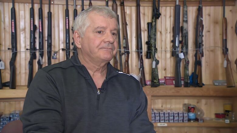 A white man sits at a counter with a display of various long guns standing against a wooden wall behind him, and ammunition visible on the shelves