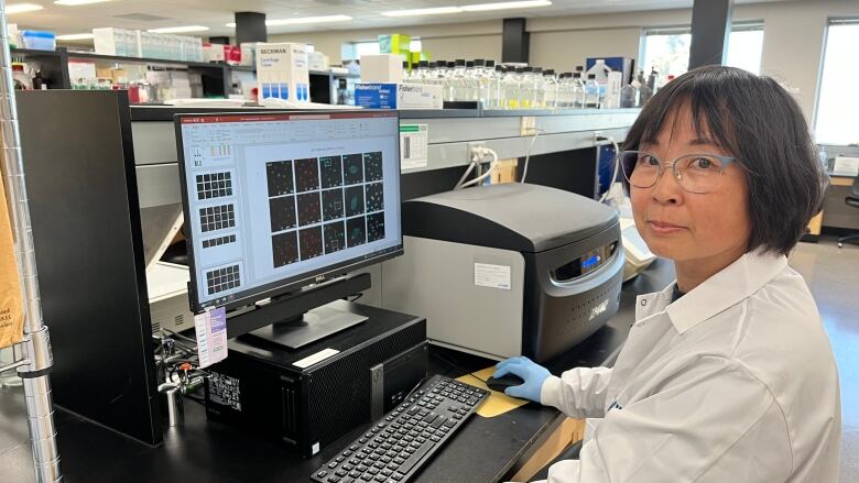 A vaccine researcher with short black hair and glasses sits at her desk in lab wearing white lab coat