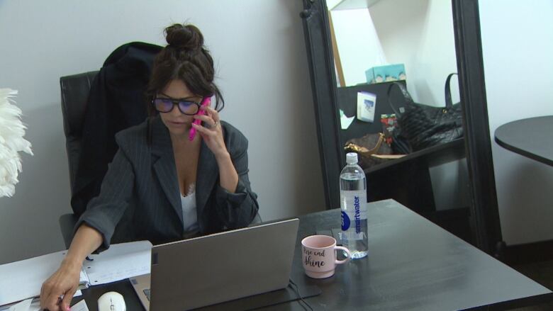 A woman at a desk holds a cell phone and looks down toward a laptop.