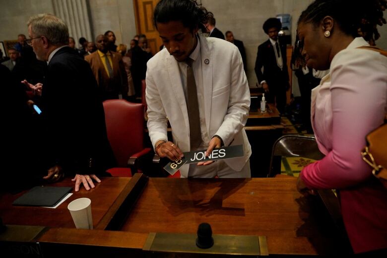 Man in white suit and brown tie removes his nameplate from his desk