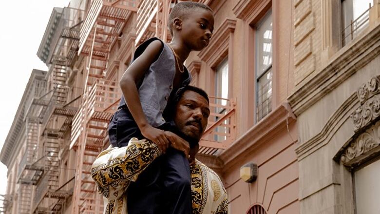 A child sits on the shoulders of a man. They are standing in front of apartment buildings. 