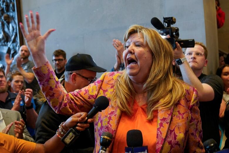 A blond woman in an orange top and pink and oraange jacket gestures as she speaks