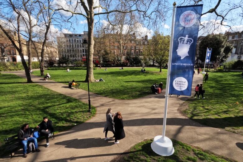 Banners fly from a pole in a park as people walk though.