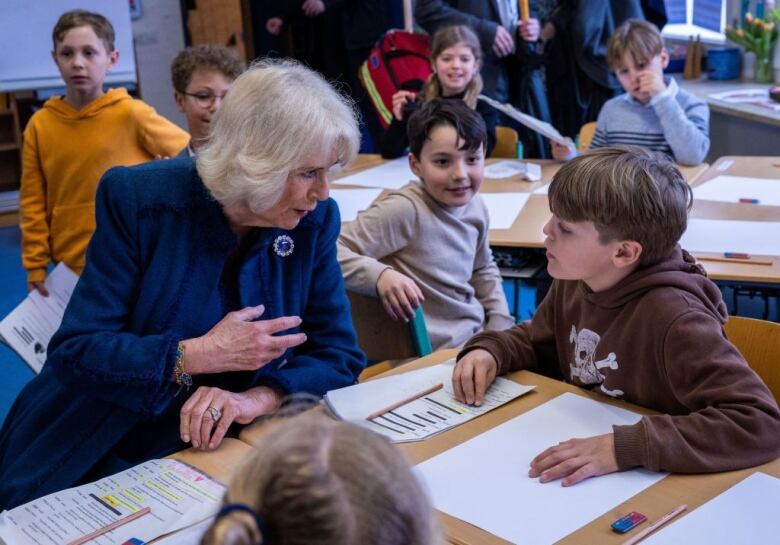 An adult sitting at a desk in a classroom talks with a child as other children look on.