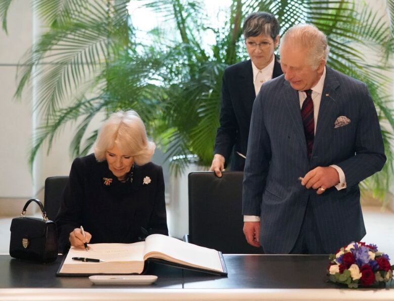 A seated person signs a guest book as two people who are standing watch.