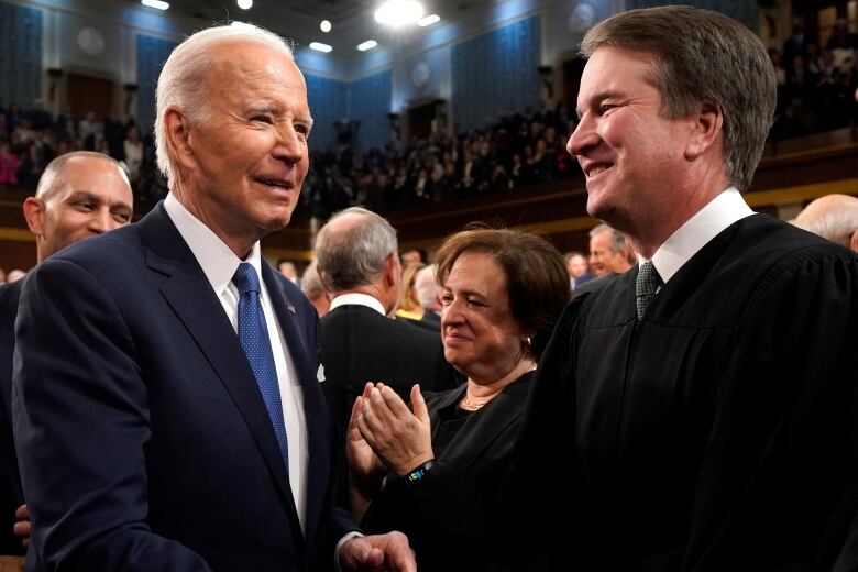 A man in a suit and tie and a man in a judicial robe and a tie are shown inside a legislative chamber.