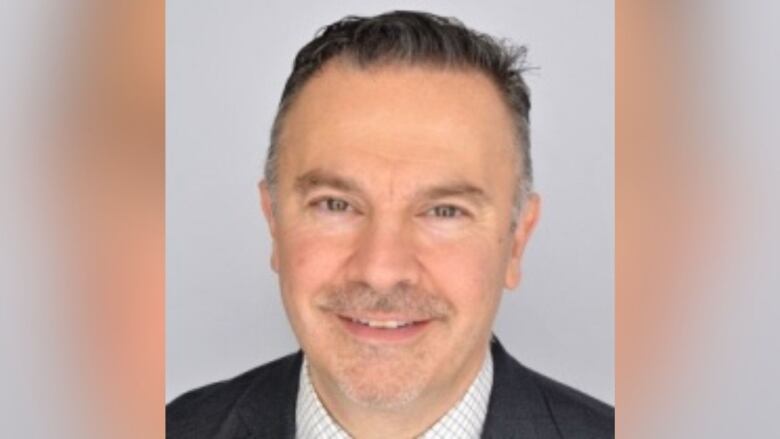 A man stands in front of a grey background. he wears a black jacket with a white shirt. He has grey salt and pepper hair and a moustache.