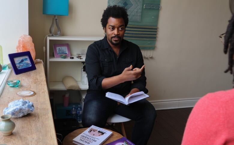 A man holding a notebook speaks with someone opposite him in an office. 