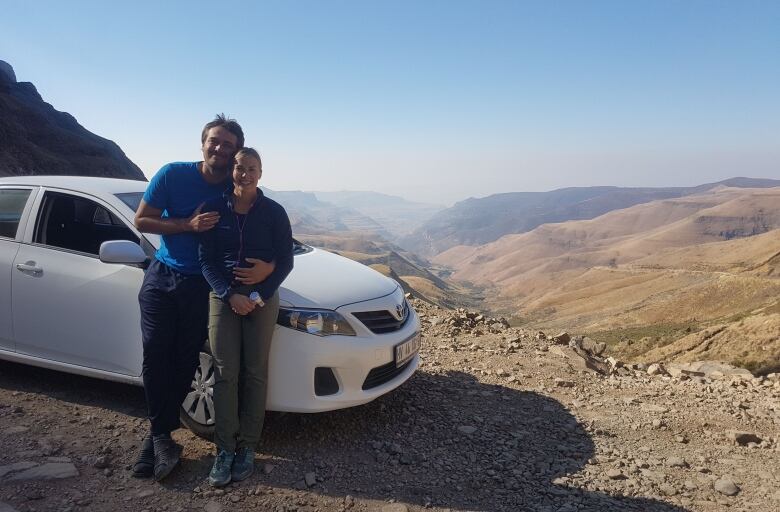 A man and woman stand in front of a white car on top of a mountain.