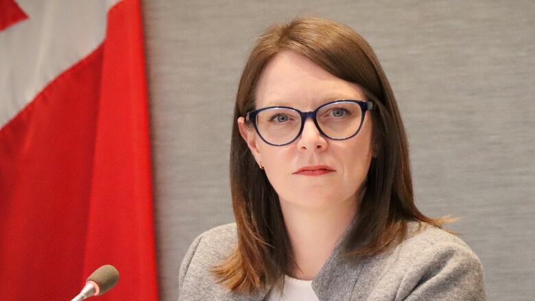 A woman with glasses and a grey jacket looks off with a Canadian flag on the wall behind her.