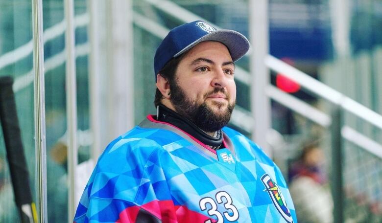 Max Denley looks forward, wearing a blue hockey jersey with a diamond pattern. He is wearing a cap and has a beard and mustache.