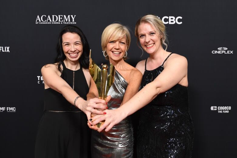 Three women in evening wear hold a gold award trophy.