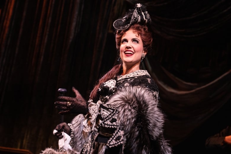 A red-haired actress wears a hat and a fur coat while in character in a Broadway show.