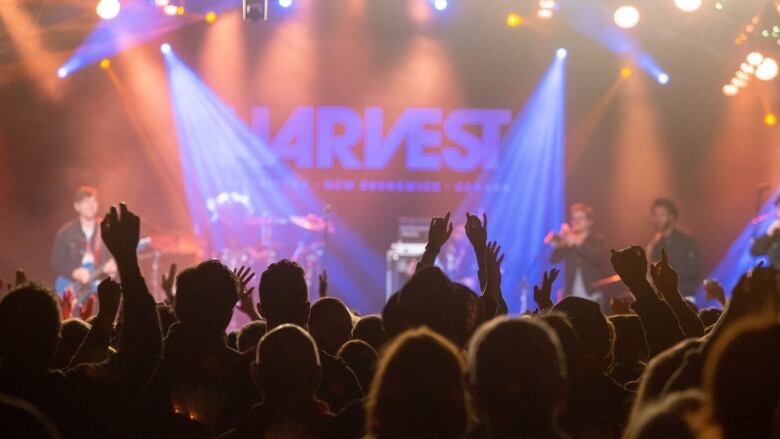 A stage lit with purple light, silhouettes of people with arms up in the foreground.