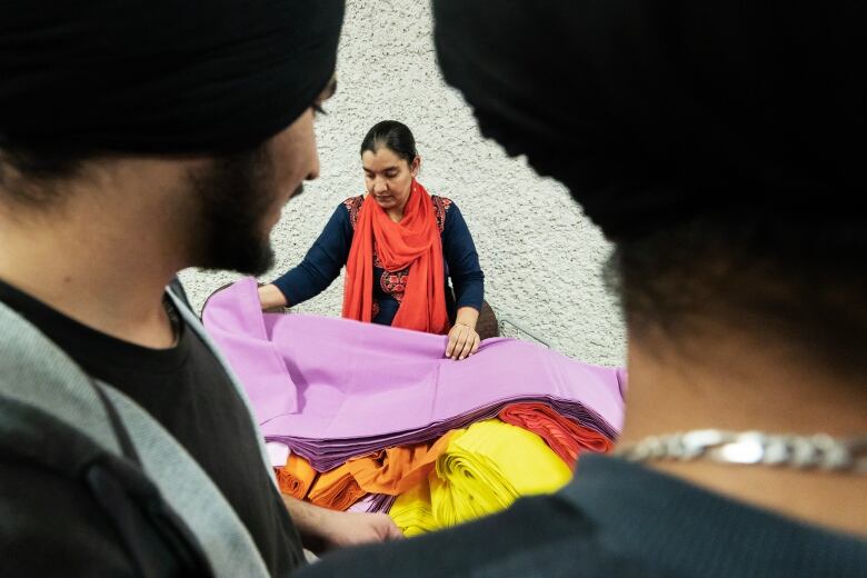 Woman  wearing red scarf around her neck and navy long sleeve sits pink fabric on top of a bundle of multi-coloured fabric. She is framed by two men wearing black turbans.