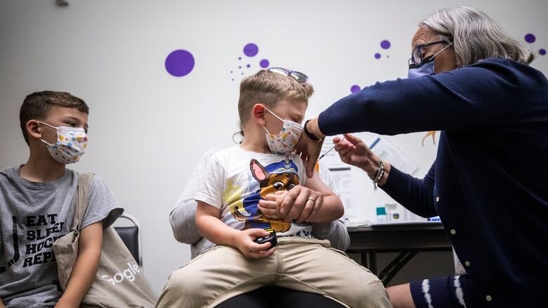 A nurse wearing a mask holds a syringe into a young boy's arm as he wears a mask and sits on his mother's lap, while another boy wearing a mask watches