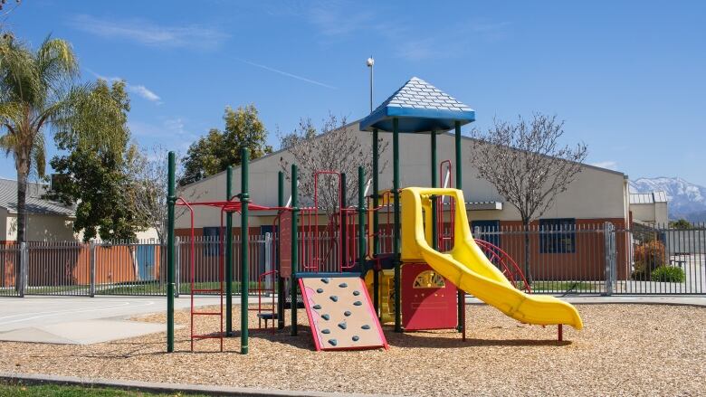 A colorful playground with ladders and slides. In the background is a brick building, trees and mountains. 