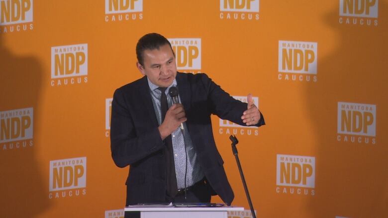 A man holding a microphone in his right hand gestures with his other hand while standing in front of an orange backdrop covered in white logos, reading, 'Manitoba NDP caucus.'