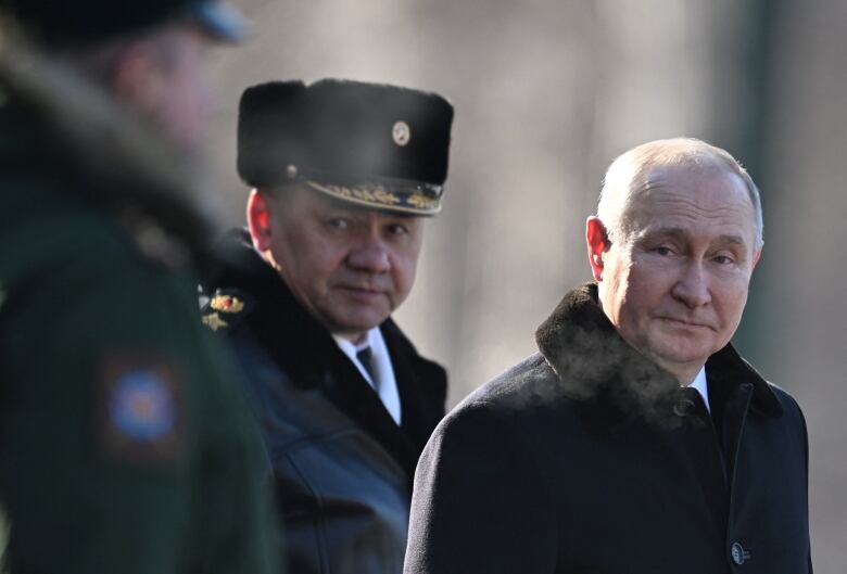 A man with grey hair and a black fur-collared coat walks outside next to an officer wearing a black fur cap with gold details.